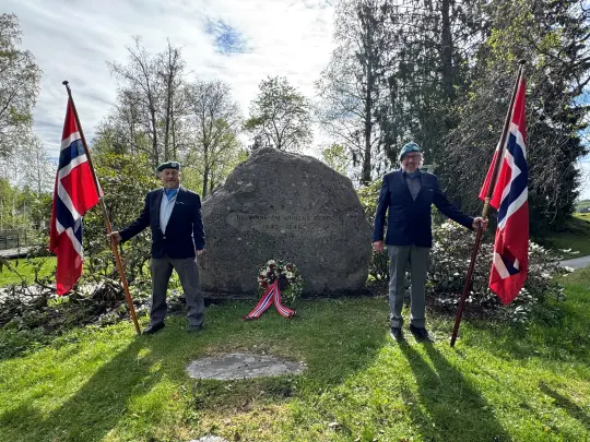 To veteraner med hvert sitt flagg foran minnesteinen i byparken.