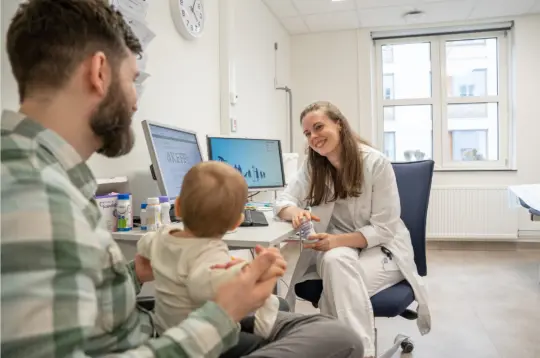Bilde av en pappa som sitter hos legen med et barn på fanget.