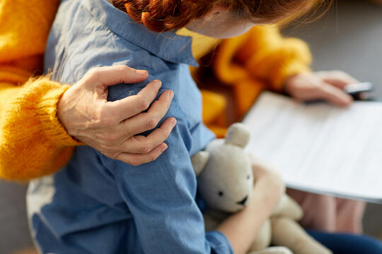 En person som holder rundt ei jente som sitter med bamsen sin. Bildet illustrerer å ta vare på et barn.
