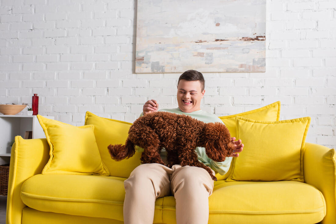 En person med utviklingshemming som sitter hjemme i sofaen og ler godt når han leker med hunden sin.